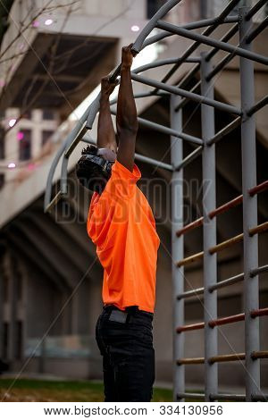 African-american Man In Orange T-hirt In Wireless Headphones Pulling Himself Up On Horizontal Bars O