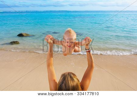 Young Happy Mother Tossing Up To Mid Air Joyful Baby Boy On White Sand Sea Beach. Outdoors Healthy C