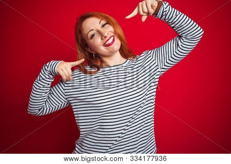 Young redhead woman wearing strapes navy shirt standing over red isolated background smiling cheerful showing and pointing with fingers teeth and mouth. Dental health concept.