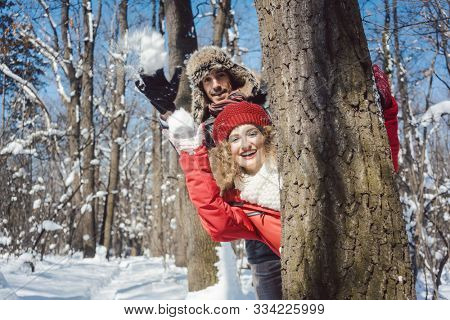 Woman and man in winter throwing snowball at the camera hiding behind a tree