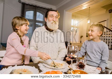 Old Handsome Grandfather With His Two Grandchildren Sitting At The Kitchen Table And Eating Pasta. L