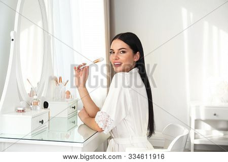 Beautiful Young Woman In Robe Doing Make Up At Dressing Table Indoors