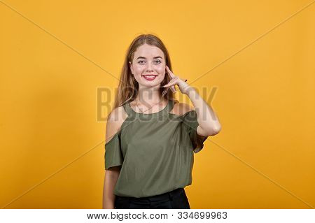 Portrait Of Cute Young Woman In Haki Orange Shirt Keeping Finger On Cheek, Smiling, Isolated On Oran