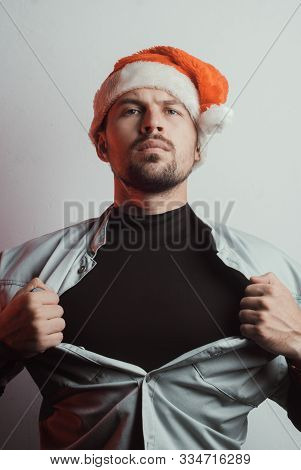A Man In A Santa Hat Rips Off His Shirt. The Hero In Black. The Concept Of Santa Claus A Superhero