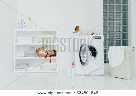 Pretty Small Child With Pigtails Lies On Shelf Of White Console With Neatly Folded Clean Towels, Pos