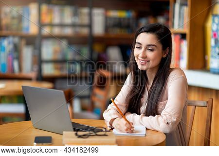 Remote Education. Happy Latin Female Student Using Laptop And Taking Notes, Sitting At Cafe Interior