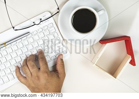 Working From Home Concept. Hand Typing On Keyboard, Spectacles, Wooden House Toy And A Cup Of Coffee