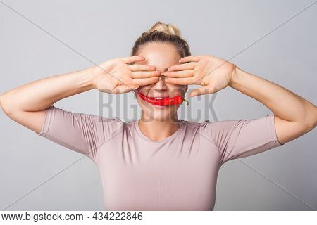 Portrait Of Young Cute Woman Covering Eyes With Hands And Biting Red Pepper, Holding Chilli In Mouth