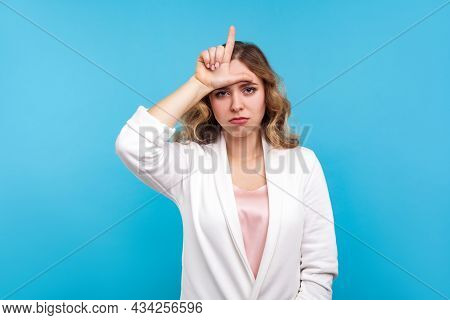Im Loser. Portrait Of Depressed Woman With Wavy Hair In White Jacket Doing Loser Gesture, L Finger S
