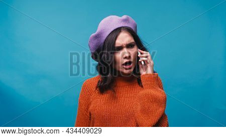 Angry Woman In Sweater And Beret Talking On Mobile Phone Isolated On Blue