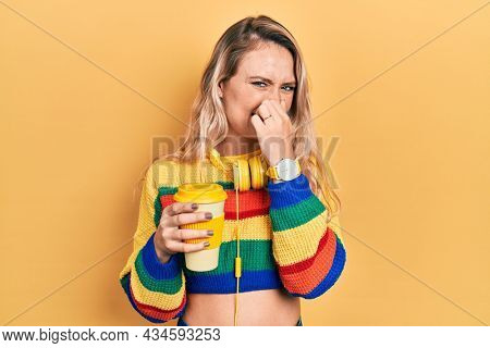 Beautiful young blonde woman drinking cup of coffee wearing headphones smelling something stinky and disgusting, intolerable smell, holding breath with fingers on nose. bad smell 