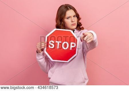 Portrait Of Serious Curly Haired Teenage Girl In Hoodie Holding Big Red Stop Road Sign And Pointing 