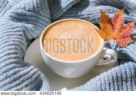 White Cup Of Morning Warming Coffee On Blue Knitted Sweater With Maple Yellow Leaves Background. Coz