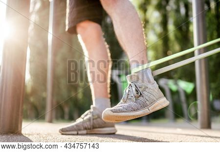 Closeup side view of legs feets of man guy training with elastic rubber band at the stadium outdoors. Adult male person during workout with additional sport equipmant in summertime