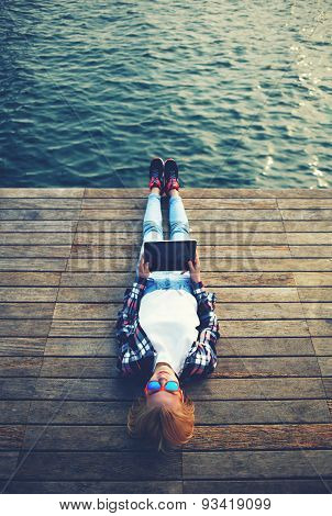 Active young stylish girl resting after a walk on a wooden pier and makes the photo on tablet