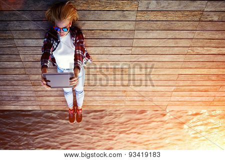 Portrait of young stylish girl is lying on a wooden pier taking self portrait on the touch tablet