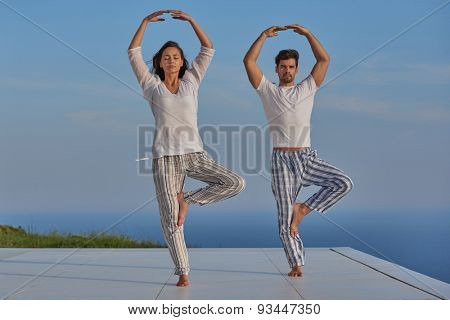 young couple practicing yoga at sunset in modern home terace with ocean and sunset in background