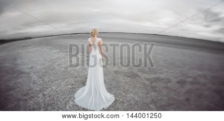 Bride outdoors in a desert looking afar. Beautiful woman in white dress full body length. Stretched wide landscape fish eye lens photo. Gray background copy space good for texting. Some fine art noise.