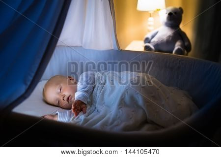 Adorable baby sleeping in blue bassinet with canopy at night. Little boy in pajamas taking a nap in dark room with crib lamp and toy bear. Bed time for kids. Bedroom and nursery interior.
