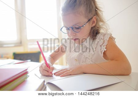 Cute little schoolgirl in glasses something diligently writes in a notebook. Girl sitting at the desk in the classroom. She is elementary school student.