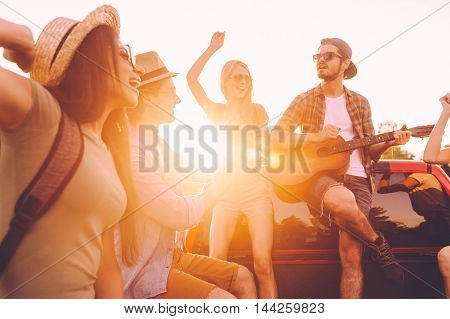 Enjoying road trip with best friends. Group of young cheerful people dancing and playing guitar while enjoying their road trip in pick-up truck together