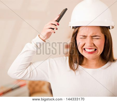 Young woman wearing construction helmet facing camera, simulating smashing mobile phone to her own head in frustration.