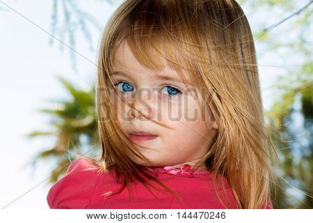A little blue-eyed girl stands with peeking through strands of her own hair as they blow in the breeze.