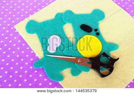 Homemade felt green animal kit, scissors on violet background with polka dots. Sewing project for children. Teddy bear patterns cut from felt. How to make a soft toy at home. Step-by-step