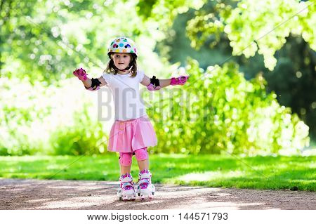Little girl learning to roller skate in sunny summer park. Child wearing protection elbow and knee pads wrist guards and safety helmet for safe roller skating ride. Active outdoor sport for kids.