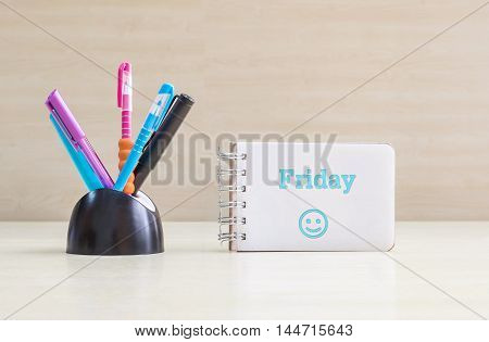 Closeup color pen with black ceramic desk tidy for pen and blue friday word in white page and happy face emotion at the note book on blurred wood desk and wood wall textured background