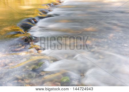 River flowing through golden and green foliage and rocks at sunlight, close up