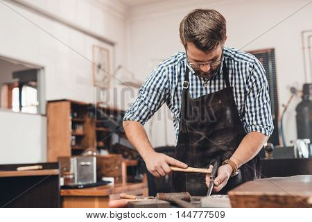 Focused jeweler in an apron using a hammer and a mandrel to shape a new ring while working at a bench in his workshop