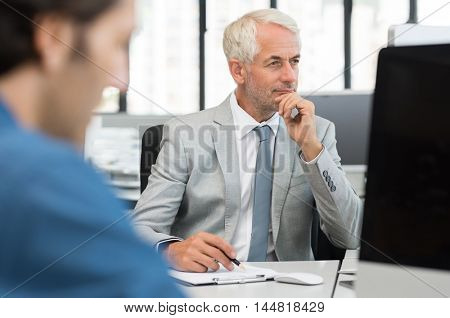 Senior businessman checking accounts in computer while tallying from report. Manager in formal reading report on desktop computer. Close up of a senior employee working with a junior employee.