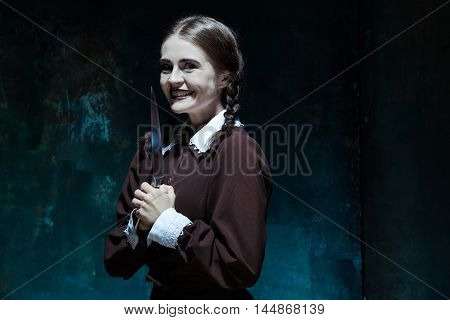 Portrait of a young girl with knife in school uniform as killer woman against school board . The image in the style of Halloween and Addams family