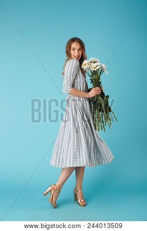 Full length image of Smiling blonde woman in dress posing sideways with flowers and looking away over blue background
