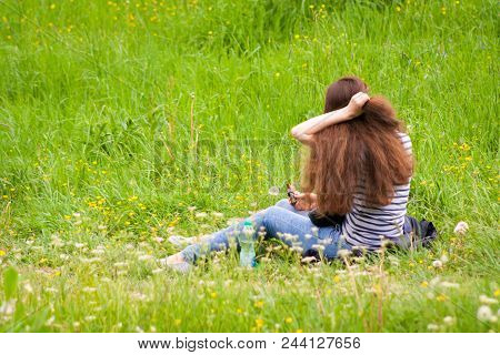 A Young Girl Sits Backwards On A Green Meadow And Improves Her Dark Hair