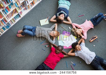 Five multiethnic children lying in circle on the floor and looking at camera. Top view of young boys and girls lying on back with drawing material. Portrait of happy smiling kids in library.