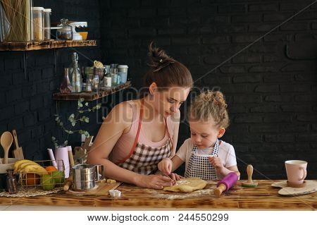 Young Mother And Daughter Prepare Cookies In Kitchen. They Are In Aprons. Little Girl And Woman Are 