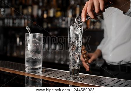 Bartender In The White Shirt Adding Ice Cubes Into An Empty Cocktail Glass On The Bar Counter On The