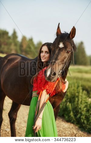 Gypsy Girl Rides A Horse In A Field In The Summer. A Woman With Long Hair Strokes And Caresses A Hor