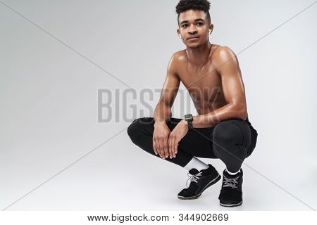 Photo of shirtless african american man using earpod?s while squatting isolated over grey background