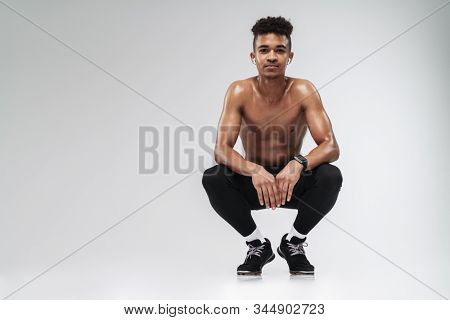 Photo of shirtless african american man using earpod?s while squatting isolated over grey background