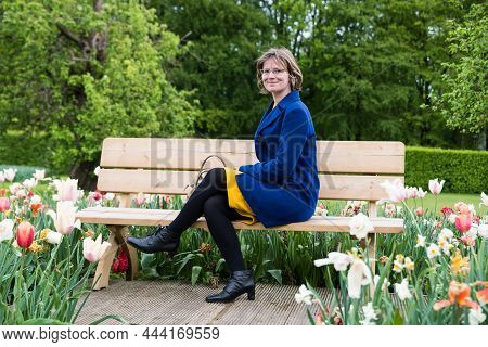 Attractive 30 Year Old Woman Sitting On A Wooden Bench, Surrounded By Blooming Flowers