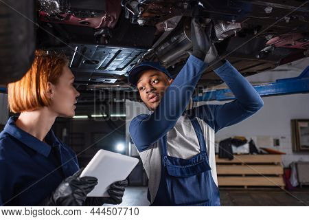 Workwoman Holding Digital Tablet Near African American Colleague Under Car In Garage.