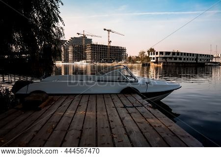 White Motor Boat Is Parked At The Pier Against The Backdrop Of Multi-storey Buildings Under Construc
