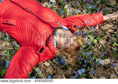 An Octogenarian Grandmother In A Red Jacket Rejoices In Spring, Lying On The Ground Covered With Blo