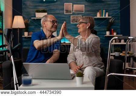 Caucasian Old People Highfive While Sitting On Sofa Using Laptop At Home. Senior Couple With Technol