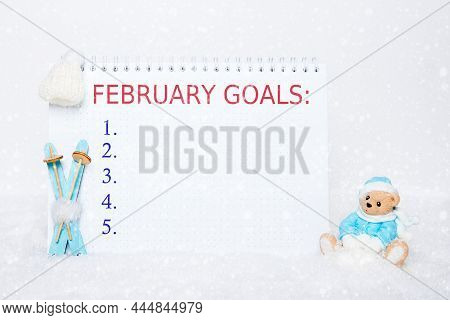 Teddy Bear In Blue Clothes Sitting On A Sled, Blue Wooden Skis, A White Hat And A Notebook With The 