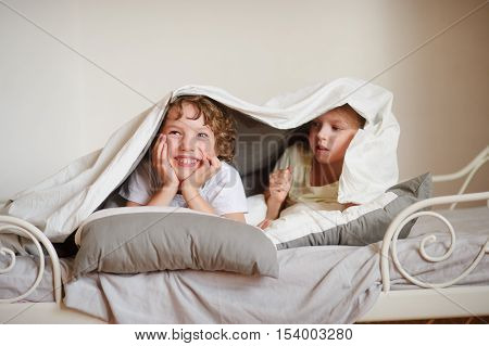 Two children brother and sister squirmy on the bed in the bedroom. They with a smile look out from under a blanket.