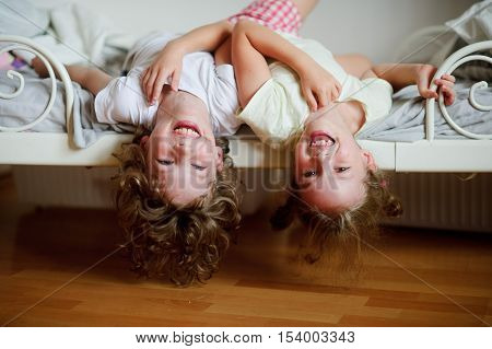 The children boy and girl naughty on the bed in the bedroom. They lie side by side their heads hanging off the bed. Children is very amused.